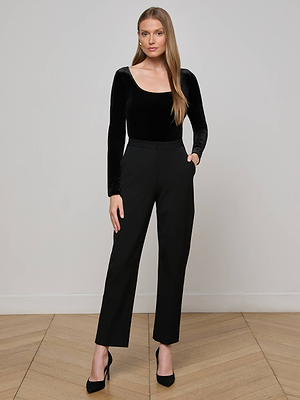 A woman with long, straight hair stands against a plain light gray wall, wearing the L'AGENCE Jelena Velvet Top, black high-waisted pants, and black heels, with one hand in her pocket and a neutral expression.