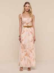 A woman stands against a plain white background, wearing the Rasha Shirred Mesh Tank—a sleeveless, floor-length dress with an abstract peach and white tie-dye print. She accessorizes with a tan belt, brown sandals, and a bracelet. Her long blonde hair frames her neutral expression.