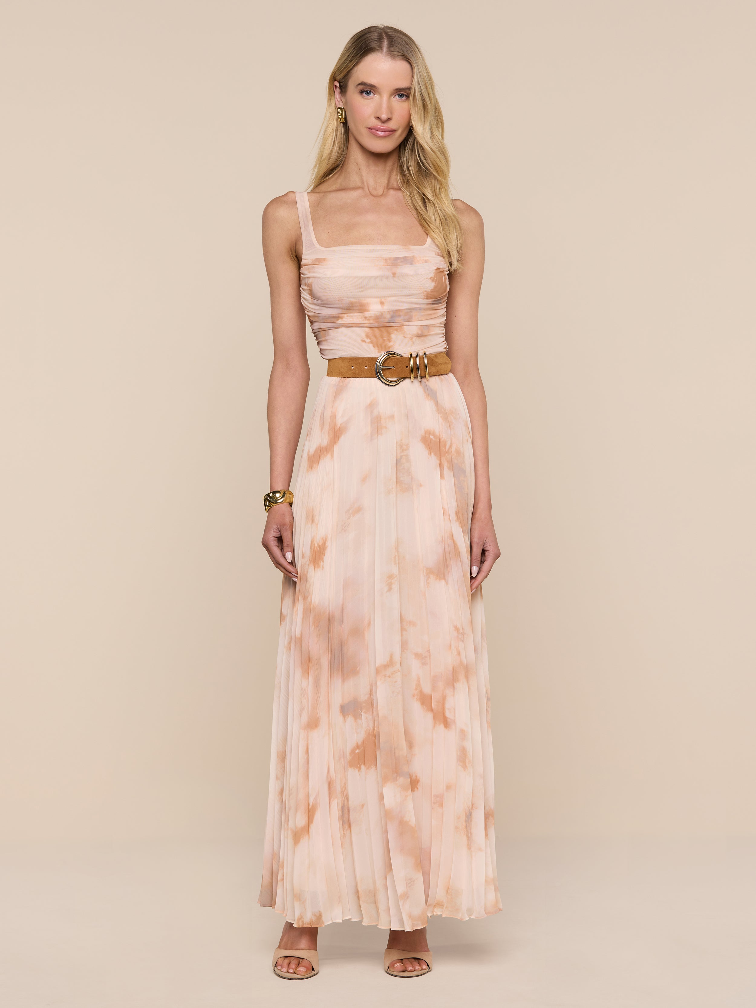 A woman stands against a plain white background, wearing the Rasha Shirred Mesh Tank—a sleeveless, floor-length dress with an abstract peach and white tie-dye print. She accessorizes with a tan belt, brown sandals, and a bracelet. Her long blonde hair frames her neutral expression.
