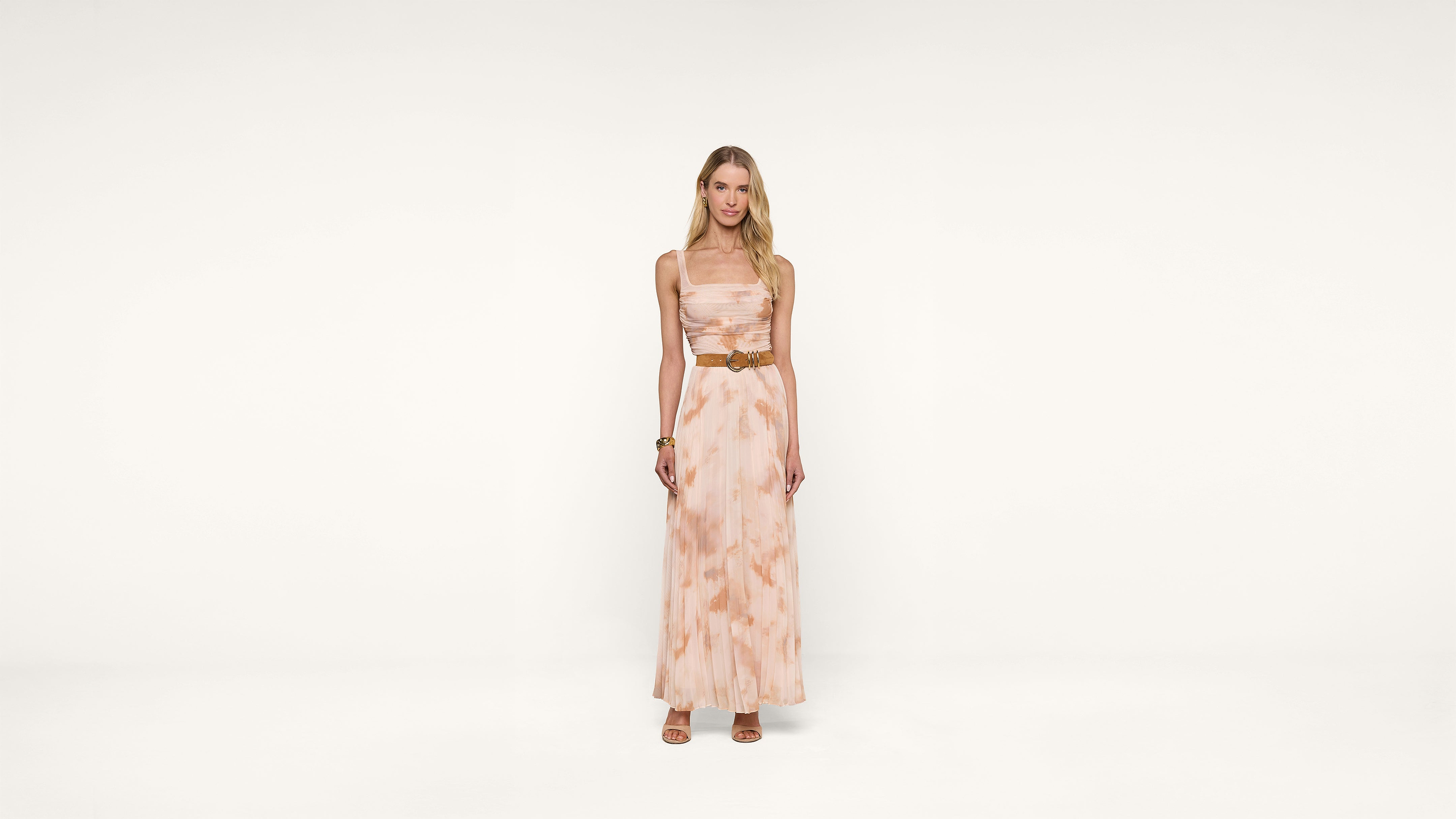 A woman stands against a plain white background, wearing a long, sleeveless, light pink and beige patterned dress with a brown belt and sandals. She has long blonde hair and a relaxed expression.