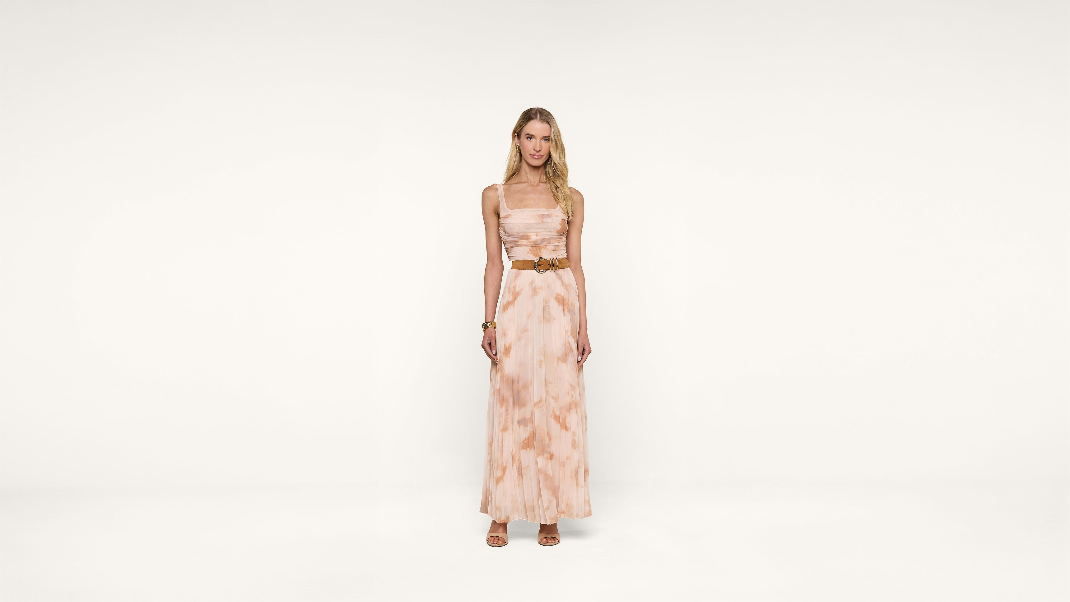 A woman stands against a plain white background, wearing a long, sleeveless, light pink and beige patterned dress with a brown belt and sandals. She has long blonde hair and a relaxed expression.