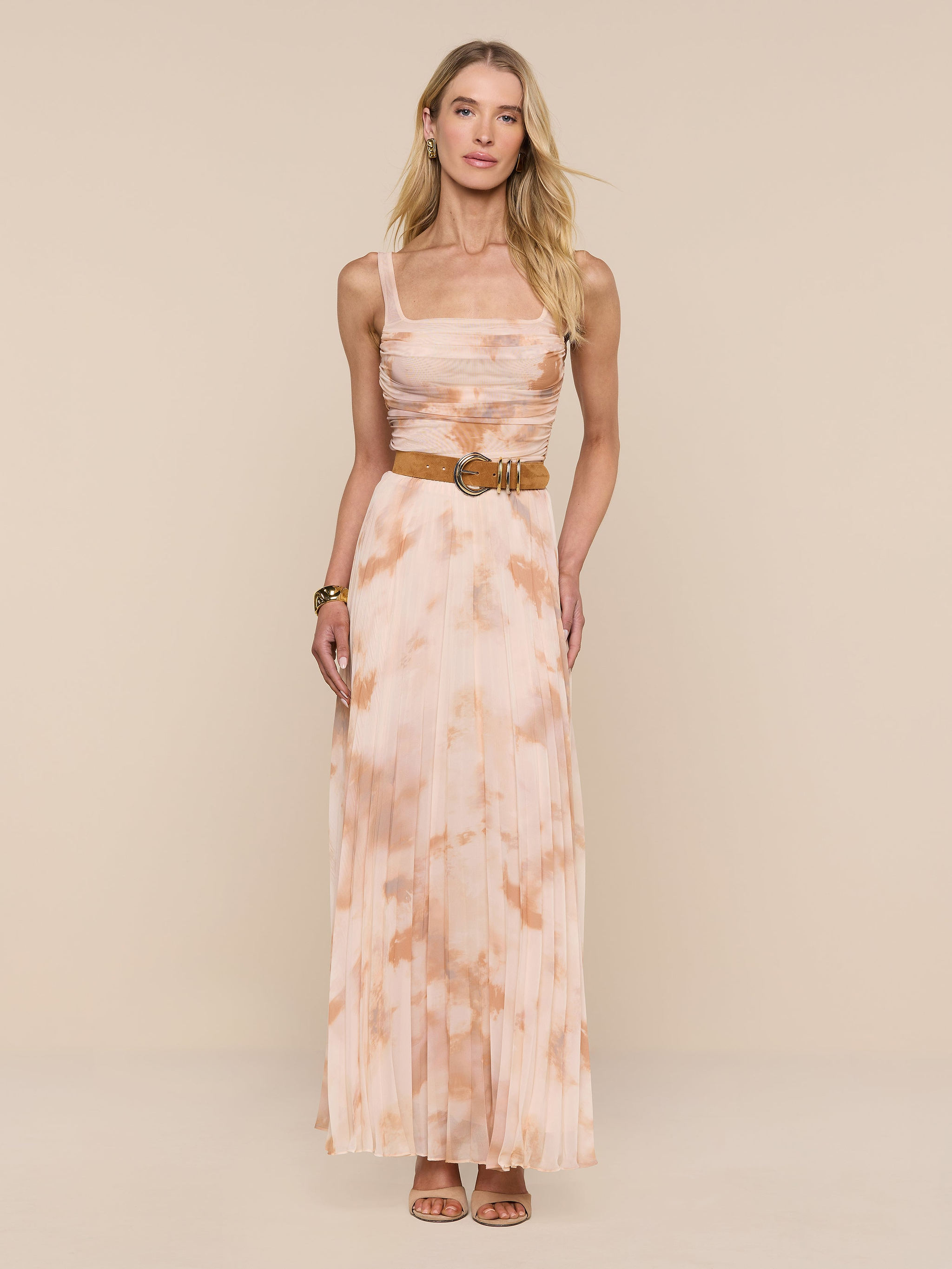 A woman stands against a white background wearing the Rasha Shirred Mesh Tank styled as a long, sleeveless dress with an abstract light pink and beige print, paired with a belt and heeled sandals.