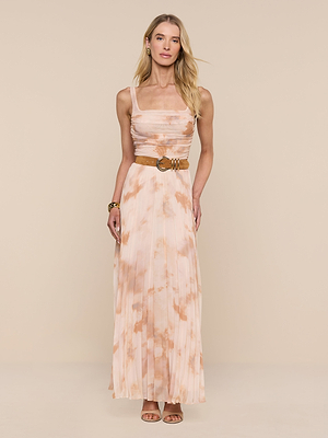 A woman with long blonde hair and a neutral expression wears the Rasha Shirred Mesh Tank in a sleeveless, peach and white abstract print maxi style, paired with a brown belt, gold bracelet, and tan sandals against a beige background.