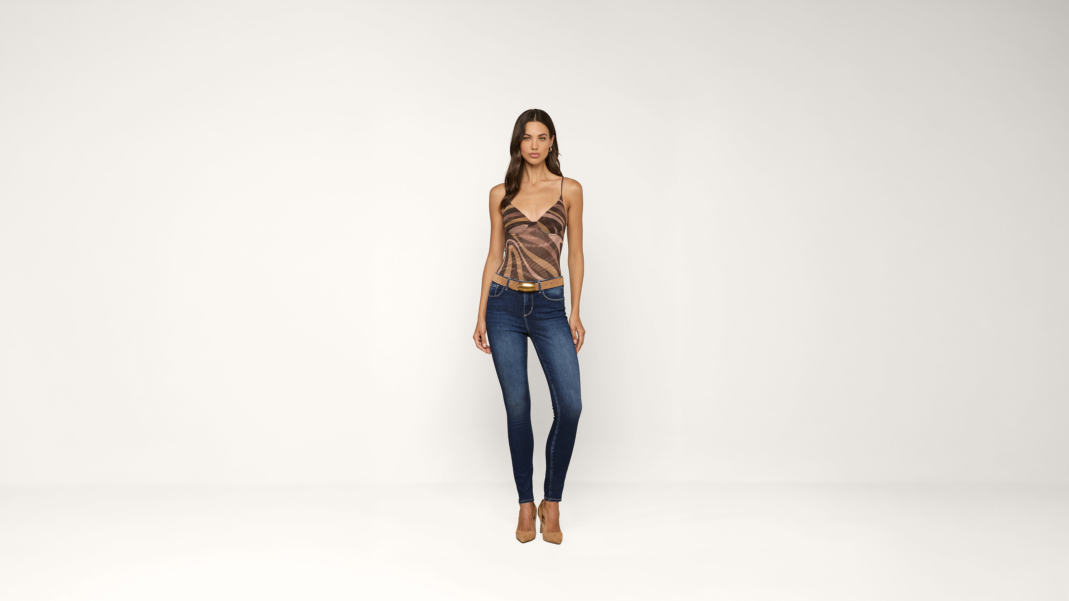 Woman standing against a plain white background, wearing a patterned sleeveless top, blue skinny jeans, and beige high-heeled shoes, with her arms relaxed at her sides.