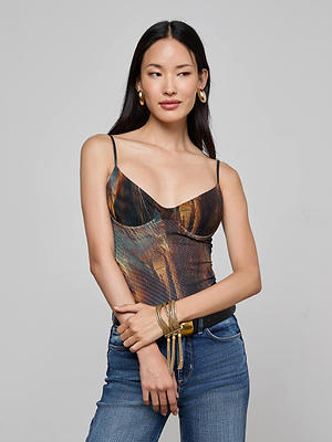 A woman with long dark hair wears the L'AGENCE Elowen Mesh Bodysuit featuring a teal feather print, paired with blue jeans and gold accessories. She stands against a plain gray background, gazing to the side with a relaxed expression.
