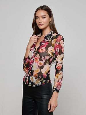 A woman with long brown hair wears the Indiana Mesh Blouse by L'AGENCE, a sheer button-down with rose floral print in red, pink, and yellow. She pairs it with black pants and stands confidently against a plain light gray background.