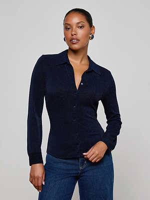 A woman with tied-back hair models the Indiana Blouse by L'AGENCE—a fitted, midnight navy button-down with subtle metallic pinstripes—paired with blue jeans. She poses confidently against a plain light gray background.