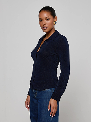 A woman stands against a plain background, wearing the L'AGENCE Indiana Blouse in midnight navy paired with blue jeans. Her hair is pulled back, and she gazes at the camera with a neutral expression.