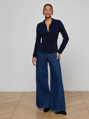 A person stands indoors by a plain wall, wearing the L'AGENCE Indiana Blouse and high-waisted, wide-leg blue jeans with black shoes. They have short hair and earrings.