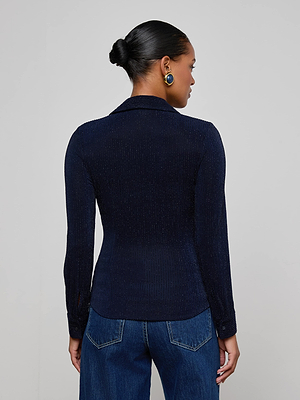 A woman with her hair in a neat bun is seen from behind, wearing the fitted Indiana Blouse by L'AGENCE—a navy button-down with metallic pinstripes—blue jeans, and round blue statement earrings.