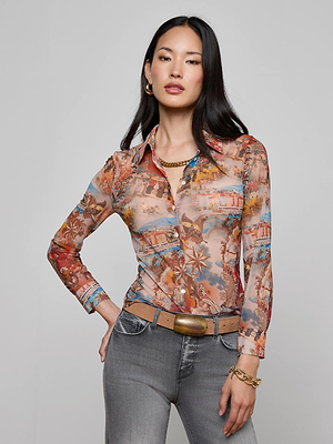 A woman with long dark hair wears the Indiana Mesh Blouse by L'AGENCE, styled with grey jeans, a gold belt, and gold jewelry, posing with one hand in her pocket against a plain light background.