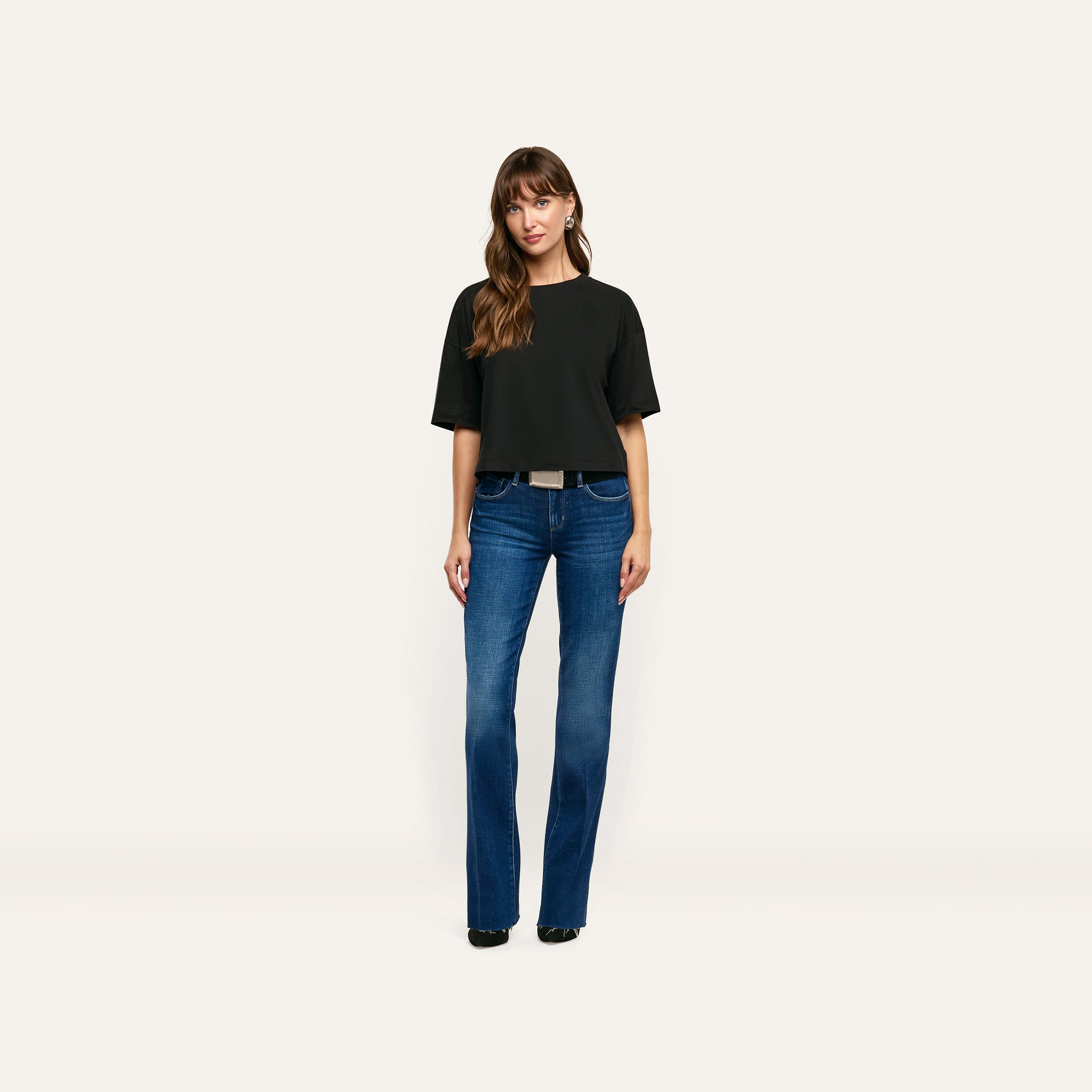 A woman wears a black relaxed-fit Daria Oversized Cropped Tee made of Supima cotton with blue bootcut jeans and a belt.