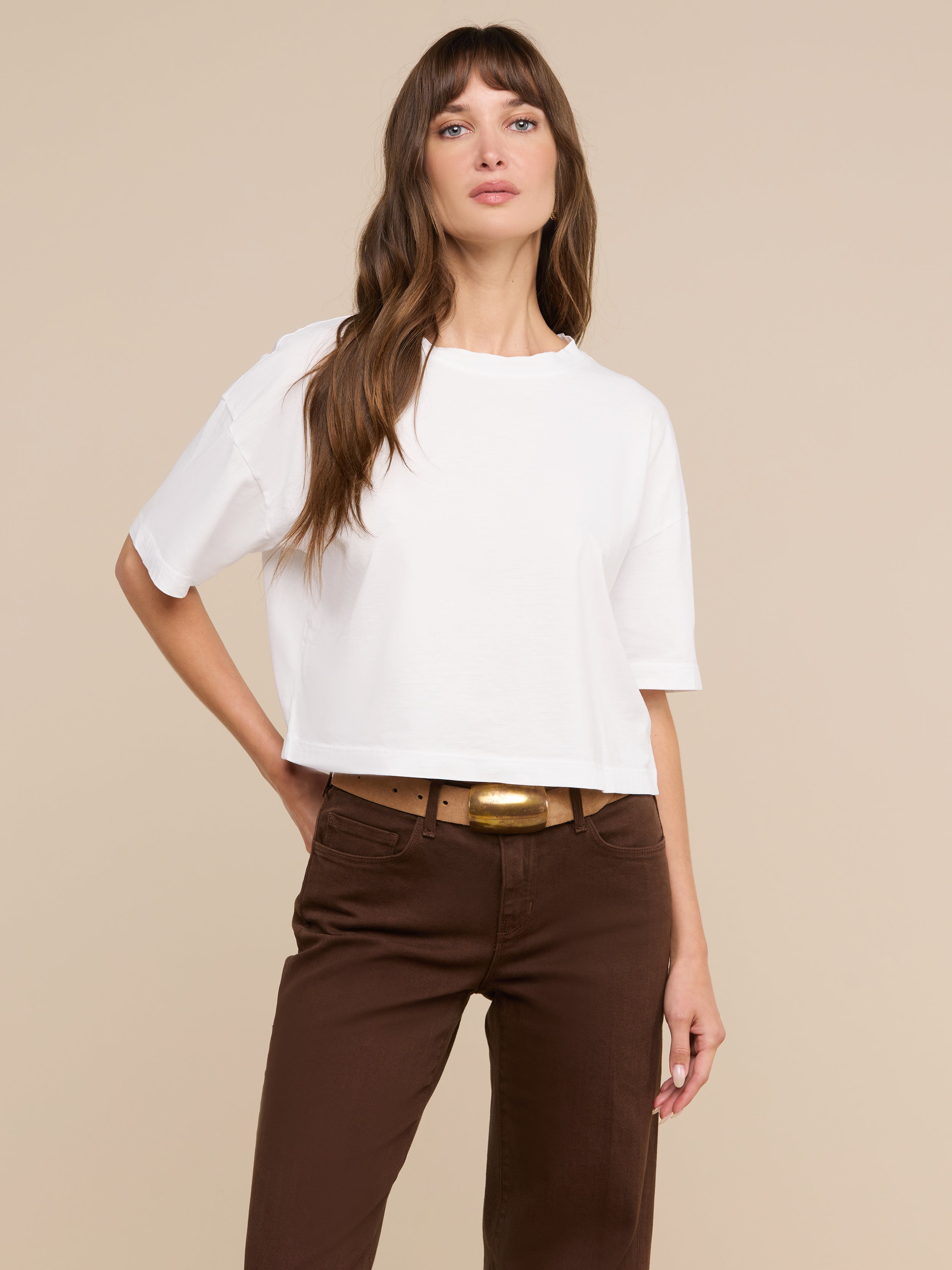 Una mujer lleva la camiseta corta oversize «Daria» en color blanco, combinada con unos pantalones marrones y un cinturón ancho, y posa ante un fondo beige con una mano en la cadera y la mirada ligeramente desviada hacia un lado.
