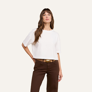 A woman wearing the Daria Oversized Cropped Tee in white, crafted from soft Supima cotton, paired with brown pants, poses with one hand in her pocket and the other relaxed by her side against a plain light background.