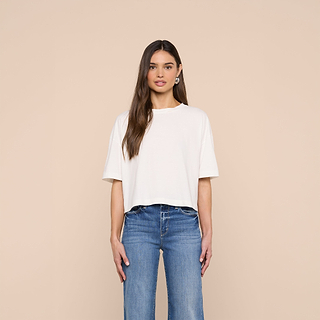 A woman stands facing forward against a beige background, wearing the Daria Oversized Cropped Tee in white—crafted from soft Supima cotton—and blue jeans. She has long brown hair, light makeup, and looks directly at the camera with a neutral expression.