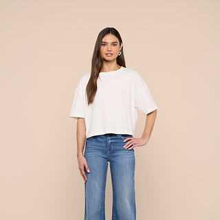 A person stands against a beige background, wearing the Daria Oversized Cropped Tee in white, crafted from soft Supima cotton, paired with blue high-waisted jeans. Their left hand rests on their hip, and they have long, straight brown hair.