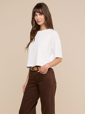 Eine Frau mit langen braunen Haaren steht vor einem beigen Hintergrund. Sie trägt das weiße „Daria Oversized Cropped Tee“ und eine braune Hose mit hoher Taille und Gürtel und blickt mit einem leichten Lächeln in die Kamera.
