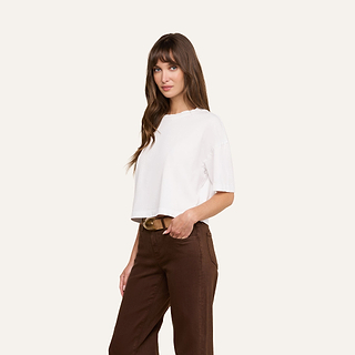 A woman with long brown hair wears the Daria Oversized Cropped Tee, styled with brown pants and standing with one hand in her pocket against a plain light background.