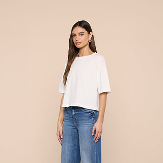 A woman with long brown hair wears the Daria Oversized Cropped Tee, a relaxed fit white top made from Supima cotton, paired with blue jeans. She stands against a plain beige background, gazing slightly to the side with a neutral expression.