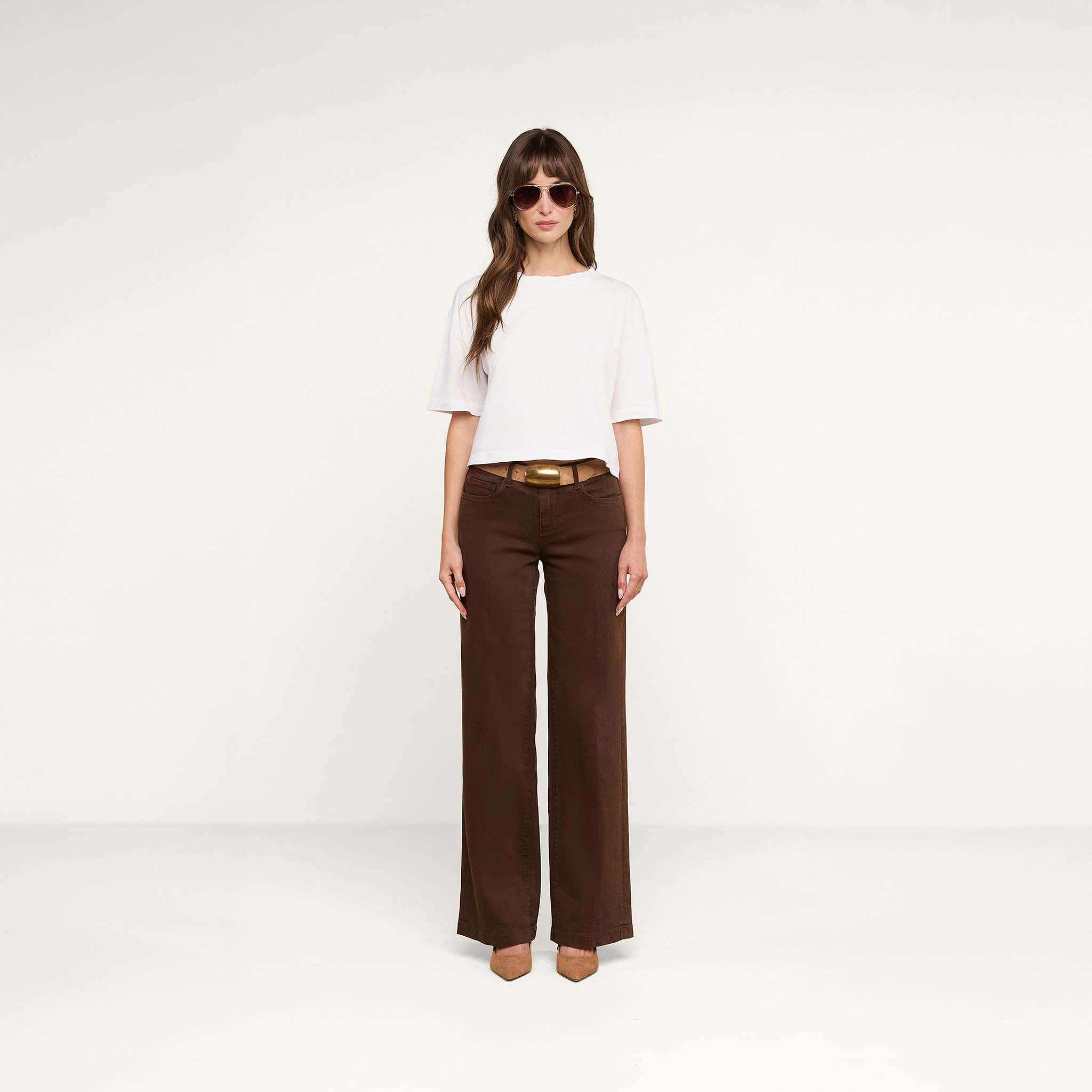 Una mujer posa ante un fondo beige con la camiseta corta oversize «Daria», confeccionada en suave algodón Supima blanco, combinada con unos pantalones anchos marrones con cinturón, zapatos de tacón marrones y gafas de sol.