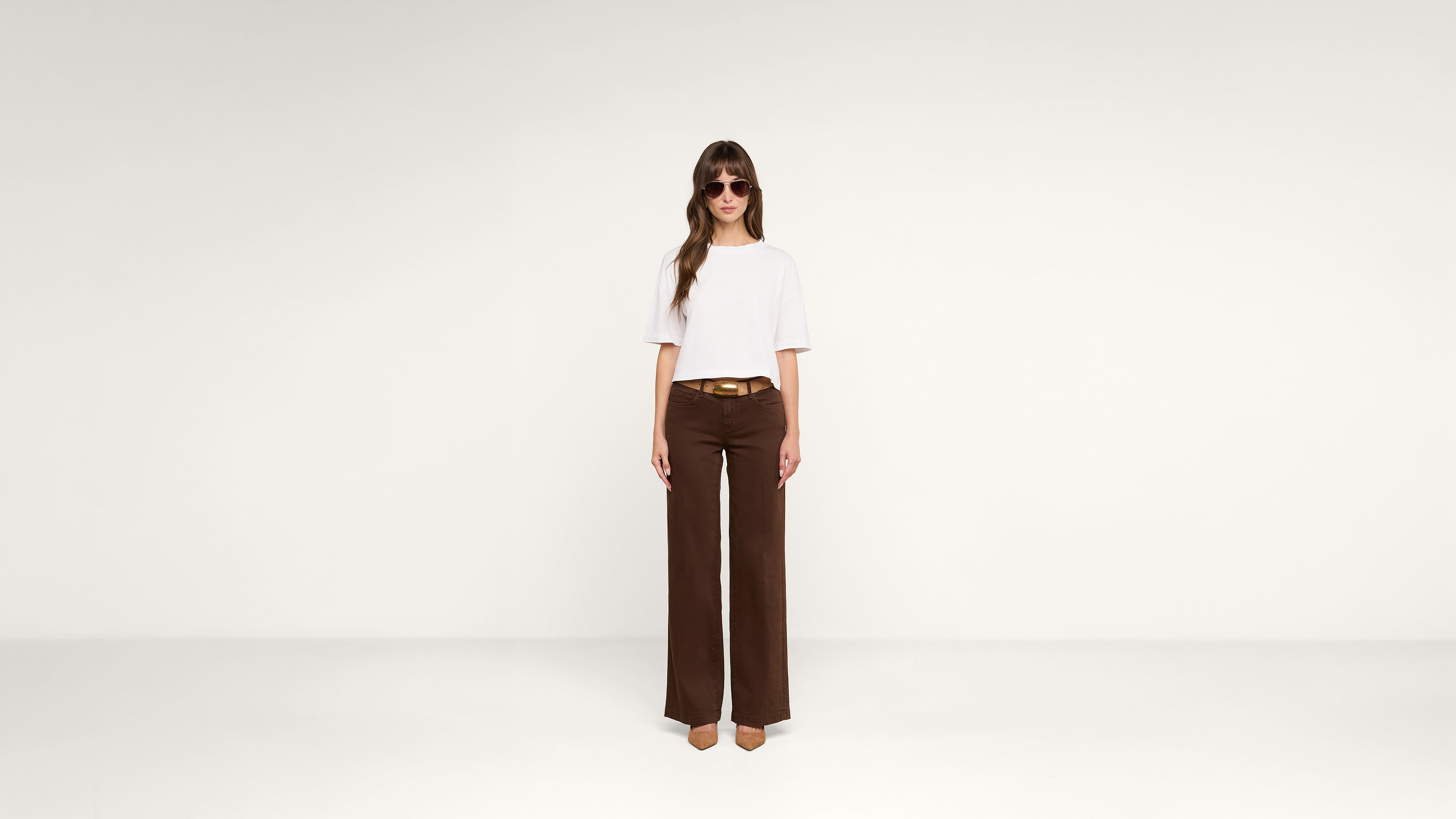 A woman wearing sunglasses, a white T-shirt, brown wide-leg pants, and tan heels stands against a plain white background.