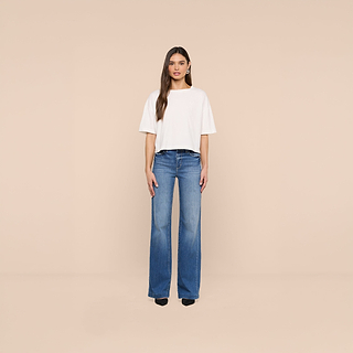 A woman stands facing forward against a beige background, wearing the Daria Oversized Cropped Tee in white crafted from soft Supima cotton, paired with blue high-waisted wide-leg jeans and black shoes.