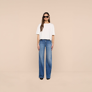 A person wearing sunglasses, the Daria Oversized Cropped Tee in white, and high-waisted blue wide-leg jeans stands facing forward against a plain beige background.