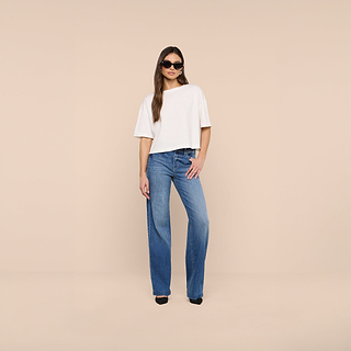 Woman wearing the Daria Oversized Cropped Tee in white—crafted from premium Supima cotton—paired with blue wide-leg jeans, black heels, and sunglasses stands against a plain beige background.