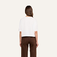 Una mujer con el pelo largo y castaño, de espaldas, lleva una camiseta corta oversize «Daria» en color blanco y unos pantalones marrones de cintura alta, todo ello sobre un fondo beige.