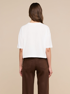 A woman with long brown hair faces away, wearing the Daria Oversized Cropped Tee in white with high-waisted brown pants against a beige background.