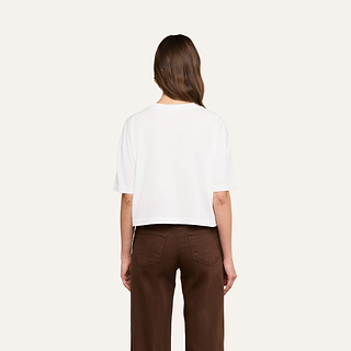 A person with long brown hair is facing away, wearing the Daria Oversized Cropped Tee in plain white Supima cotton with short sleeves and paired with brown pants against a light background.