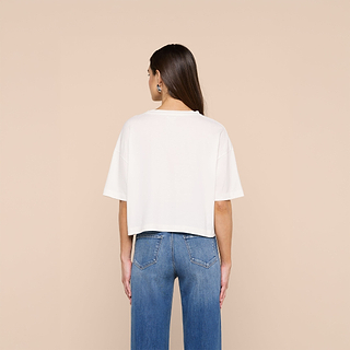 A woman with long brown hair, dressed in the Daria Oversized Cropped Tee and blue jeans, stands facing away against a beige background.