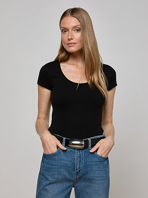 A woman with long, straight blonde hair stands against a plain light gray background, wearing L'AGENCE's Brynlee Scoopneck Tee in black and blue jeans, with her hands in her pockets.