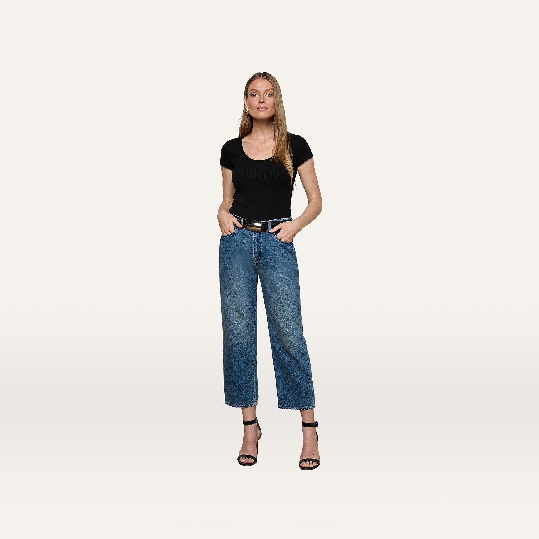 A woman stands against a plain white background, wearing the Brynlee Scoopneck Tee in black, paired with blue high-waisted jeans, a black belt, and black high-heeled sandals, with her hands in her pockets.