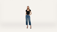 A woman stands against a plain white background, wearing a black t-shirt, blue jeans, and black high-heeled sandals. Her hands are in her pockets, and she faces forward with a neutral expression.