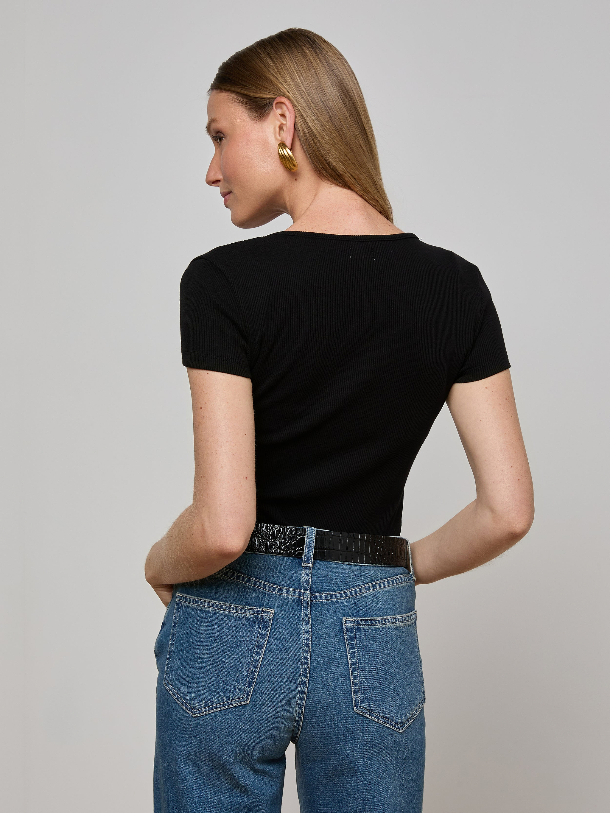 A woman with straight light brown hair, wearing the Brynlee Scoopneck Tee in black with short sleeves, blue jeans, gold earrings, and a black belt, stands with her back to the camera and her hands in her back pockets.