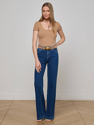A woman stands indoors against a white wall, wearing the L’AGENCE Brynlee Scoopneck Tee in beige with blue high-waisted jeans and open-toe heels. She has long hair, is smiling slightly, and poses with one hand on her hip.