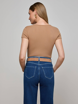A woman faces away from the camera, wearing the L'AGENCE Brynlee Scoopneck Tee in tan, paired with high-waisted blue jeans featuring waist cut-outs and accessorized with gold hoop earrings.