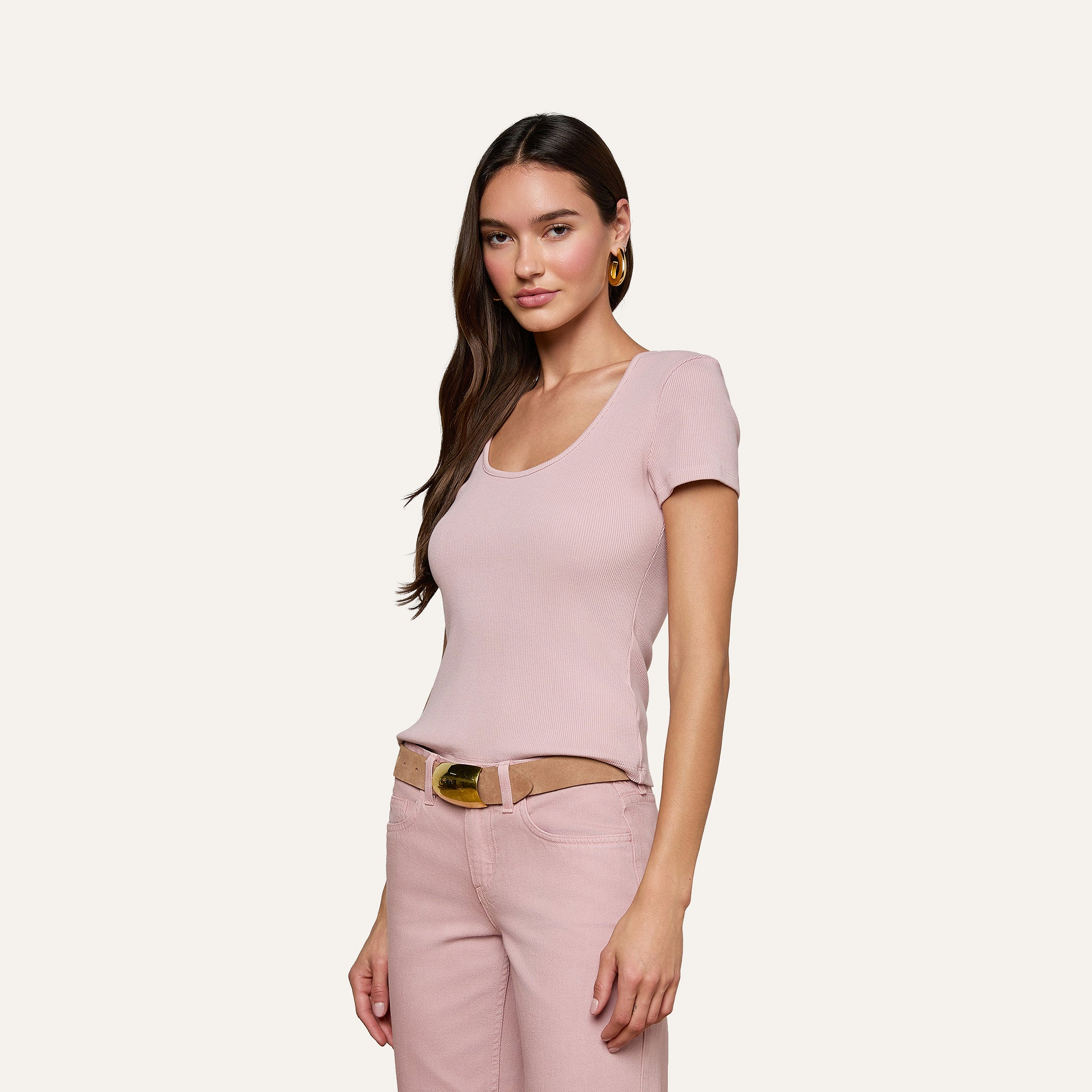 A woman with long brown hair wears the Brynlee Scoopneck Tee in light pink, paired with pink jeans and a tan belt, standing against a plain light gray background and smiling slightly at the camera.