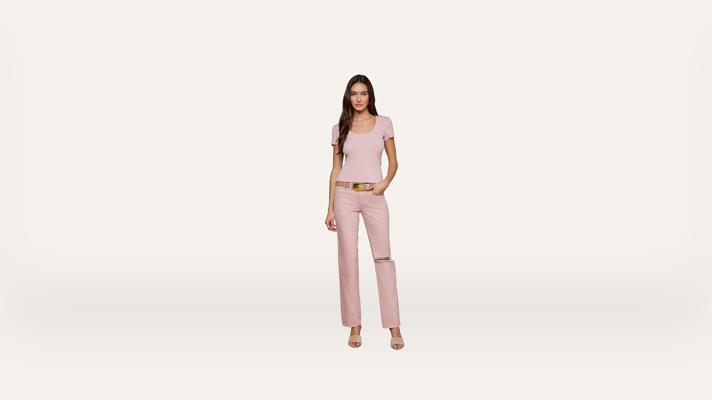 A woman stands against a plain white background, wearing a light pink t-shirt, matching pink pants with a rip at the knee, a brown belt, and open-toe heels.