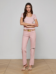 A woman with long brown hair and a gentle smile stands on a wooden floor near a light wall, wearing the Brynlee Scoopneck Tee in light pink, pink ripped-knee jeans, a beige belt, and nude heels.
