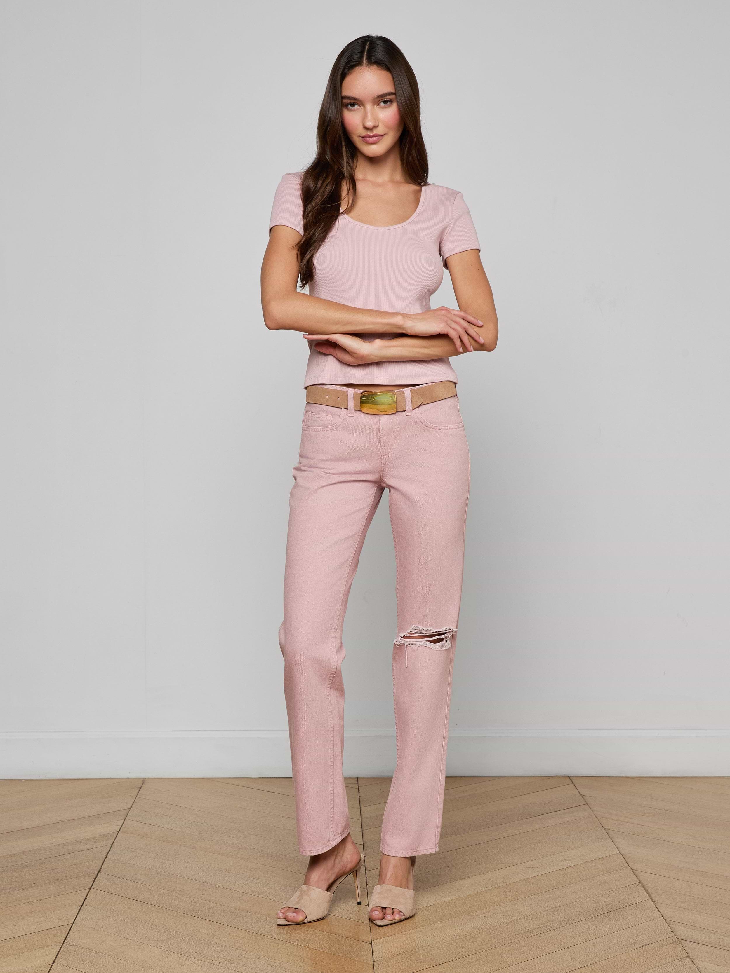 A woman with long brown hair and a gentle smile stands on a wooden floor near a light wall, wearing the Brynlee Scoopneck Tee in light pink, pink ripped-knee jeans, a beige belt, and nude heels.