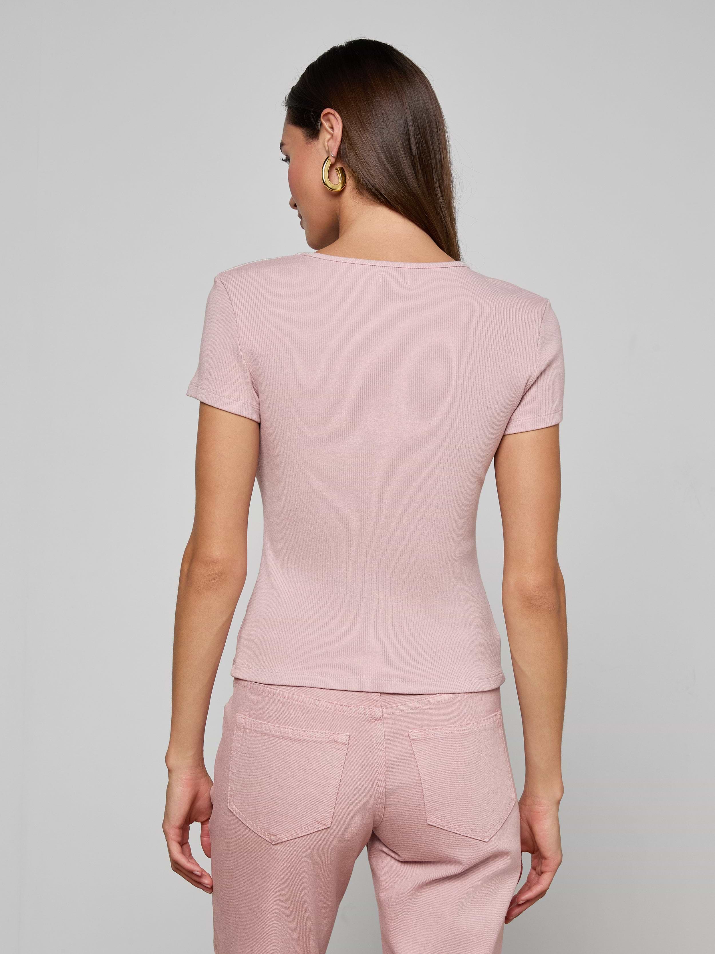 A woman with long brown hair and gold hoop earrings wears the Brynlee Scoopneck Tee in light pink and pink denim jeans, standing with her back to the camera against a plain light background.
