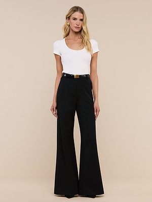 A woman with long blonde hair and a neutral expression stands against a beige background, wearing the Brynlee Scoopneck Tee in white, tucked into high-waisted black wide-leg pants with a black belt.