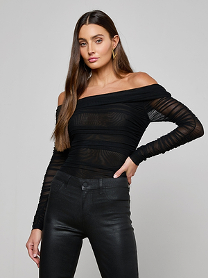 A woman with long brown hair wears the Poppie Off-The-Shoulder Bodysuit in sheer black mesh, paired with high-waisted black pants. She poses confidently against a plain light gray background, looking at the camera.