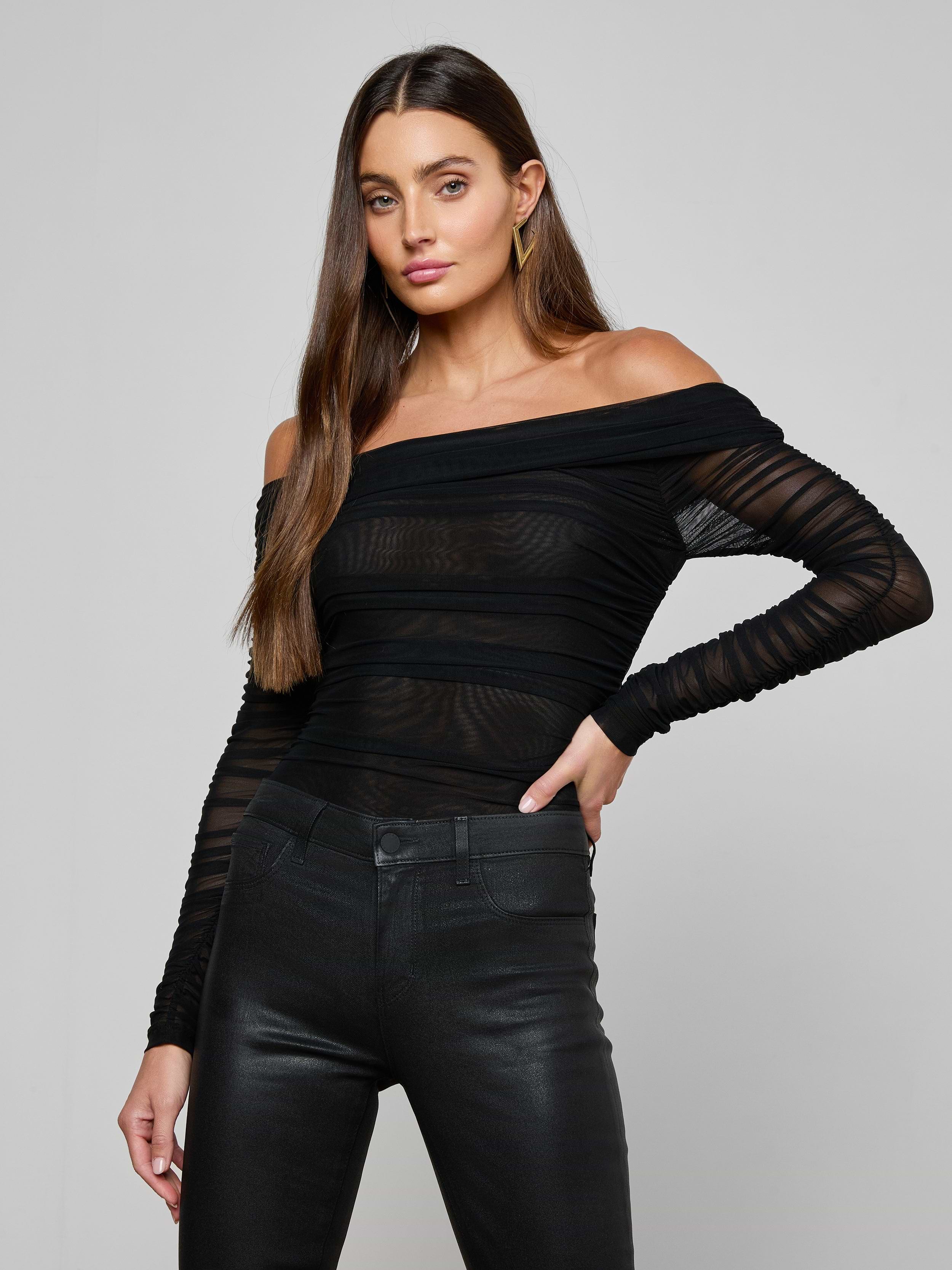 A woman with long brown hair poses with one hand on her hip against a plain light background, wearing the Poppie Off-The-Shoulder Bodysuit in black stretch mesh paired with black pants.