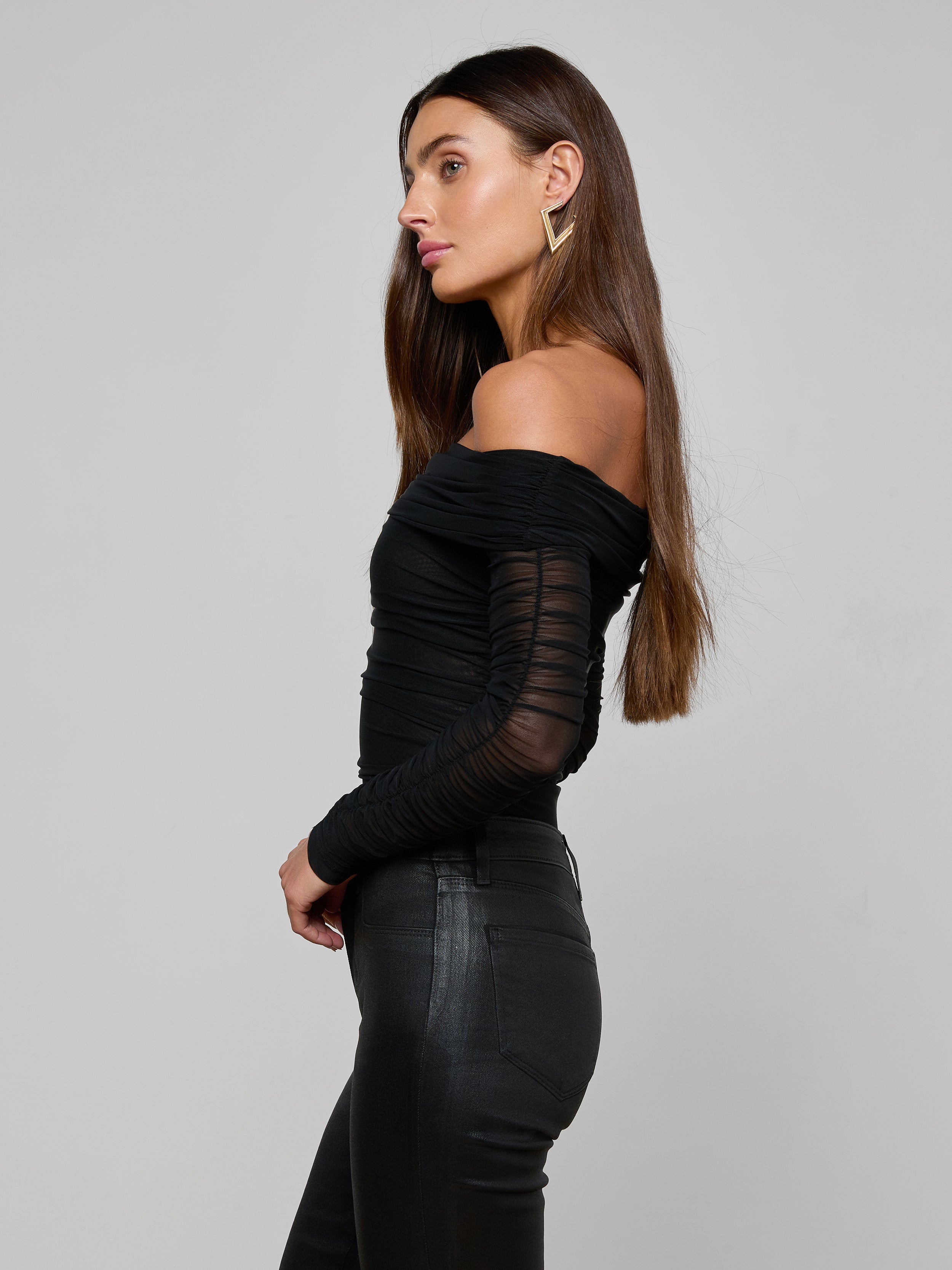 A woman in profile wearing the Poppie Off-The-Shoulder Bodysuit—with stretch mesh sleeves—paired with black fitted pants, standing against a plain, light background.