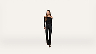 A woman with long brown hair stands against a plain white background, wearing an off-the-shoulder black top, black pants, and black heeled sandals, looking slightly to her left.