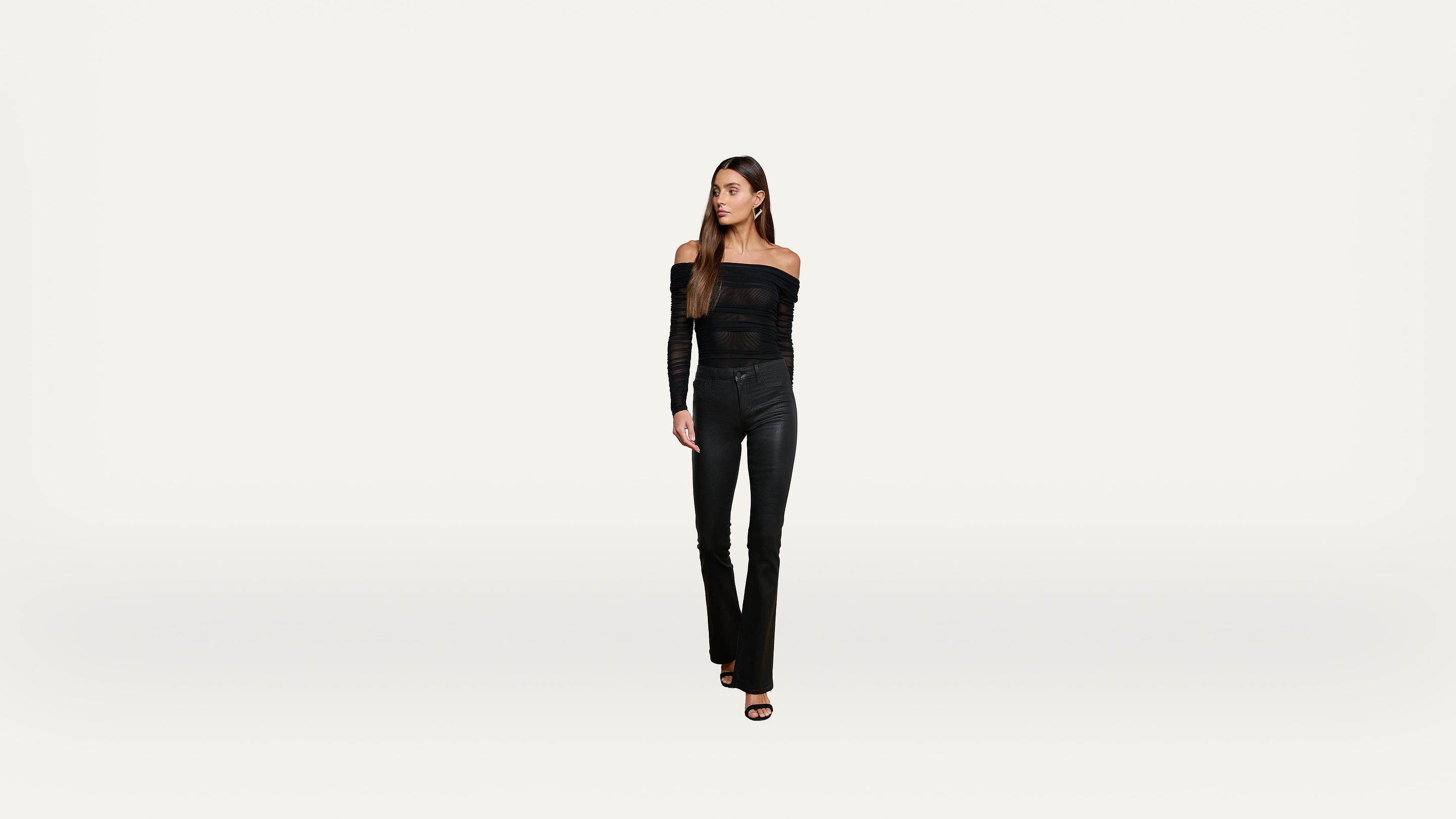 A woman with long brown hair stands against a plain white background, wearing an off-the-shoulder black top, black pants, and black heeled sandals, looking slightly to her left.