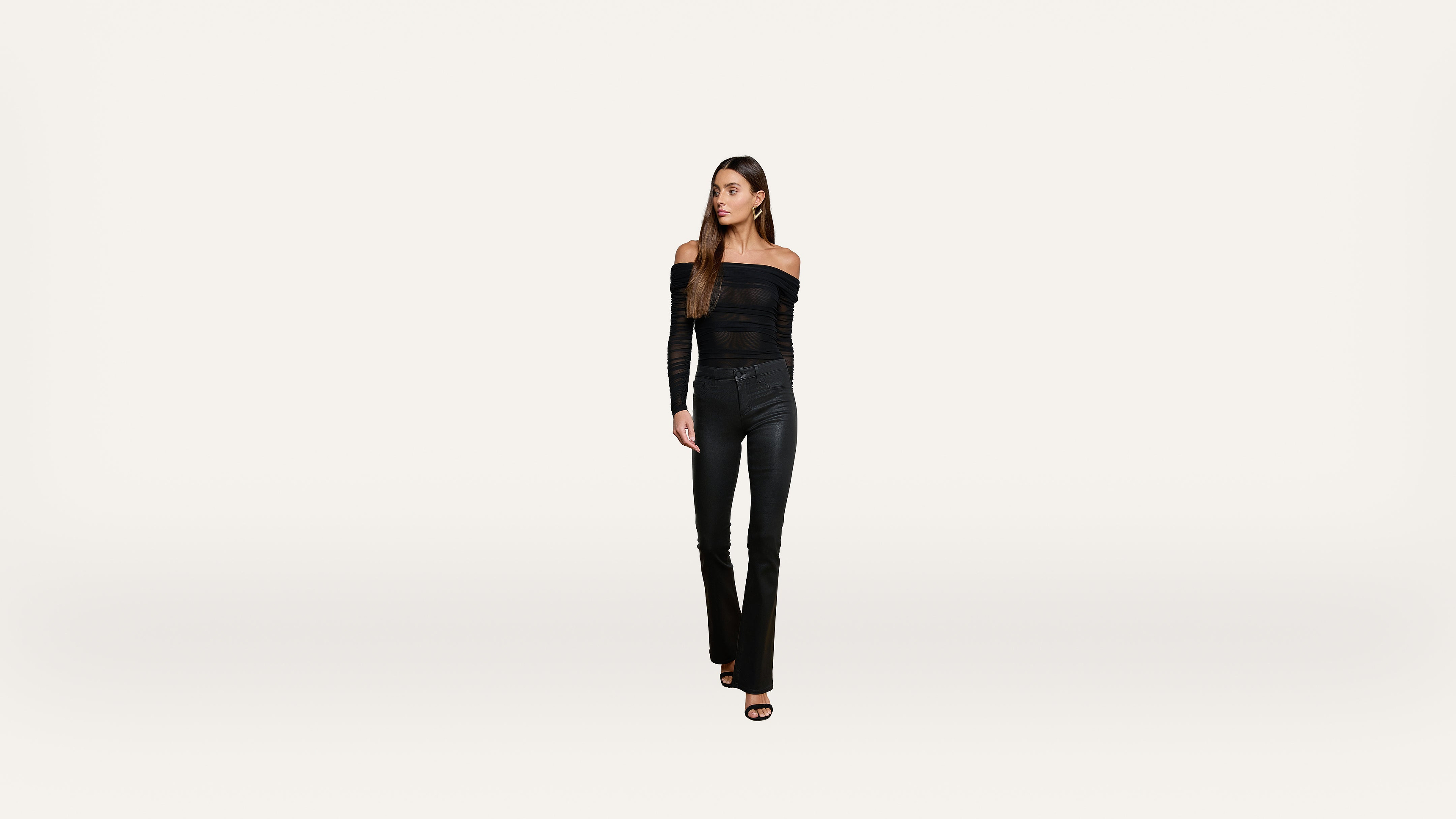 A woman with long brown hair stands against a plain white background, wearing an off-the-shoulder black top, black pants, and black heeled sandals, looking slightly to her left.